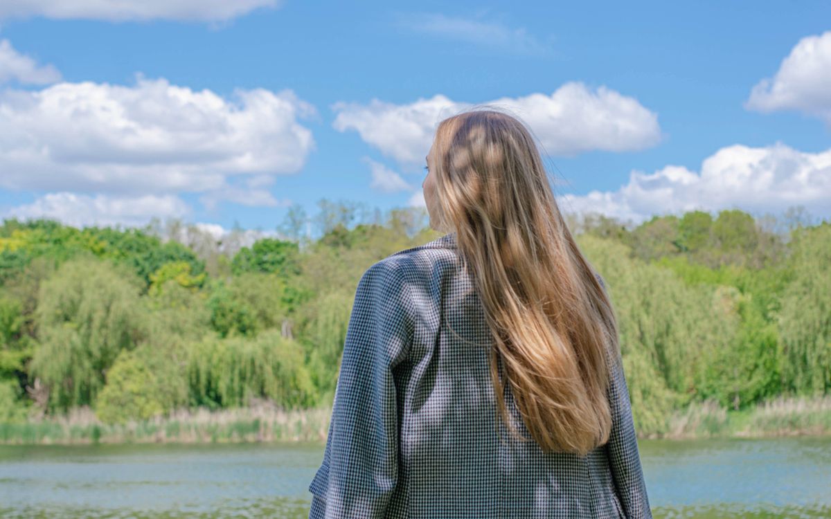 A woman with long blonde hair gazes at a tranquil lake under clear skies, surrounded by lush greenery.