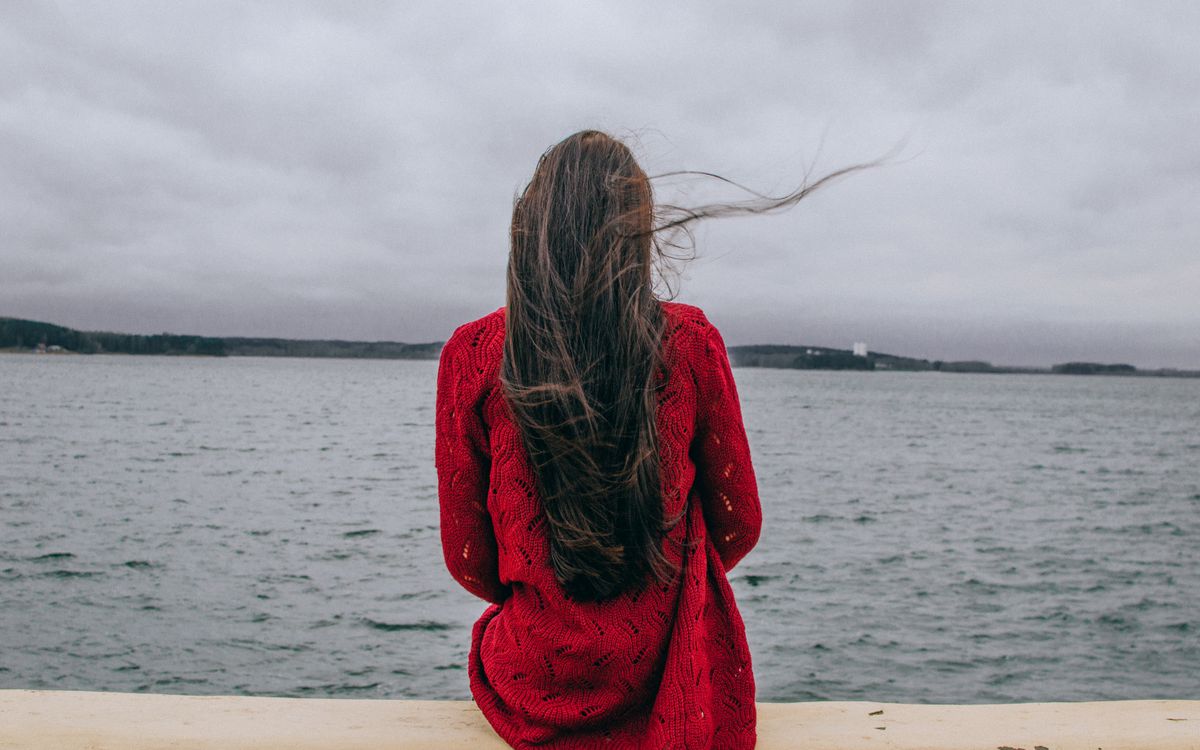 A woman in a red sweater gazing at the sea on a cloudy day.