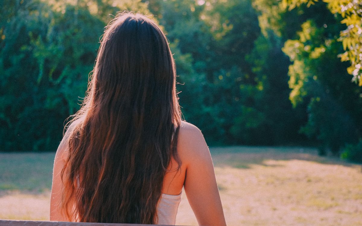 Back view of a woman with long brown hair enjoying a peaceful moment in a lush green park.