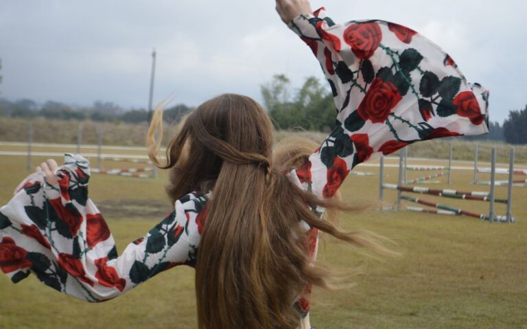 A woman with flowing brunette hair dances freely in an open field wearing a floral top.