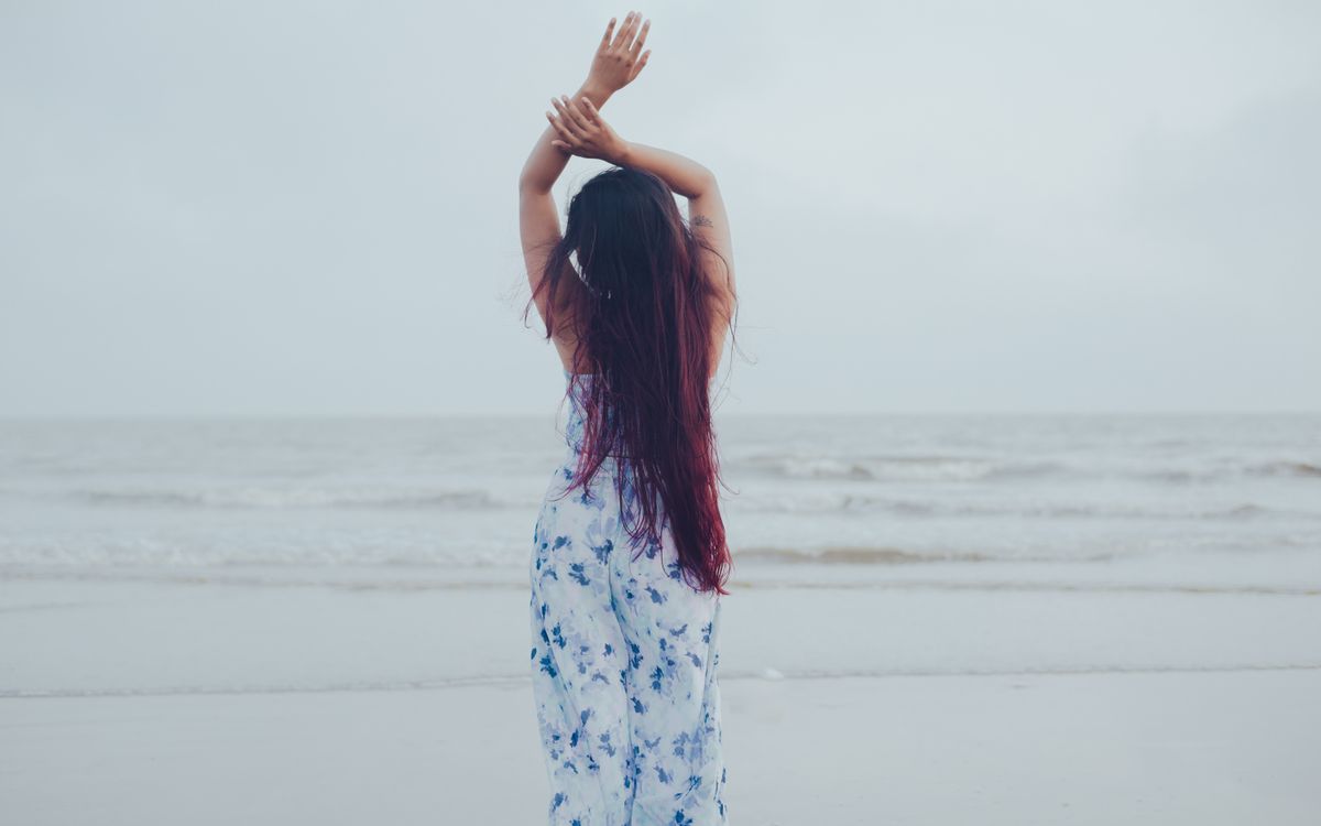 A woman with long hair in a floral dress stands on a beach, embracing the ocean breeze.