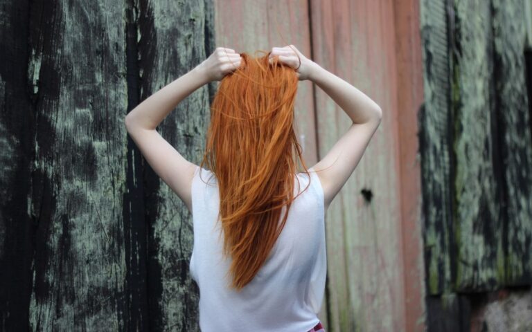 A red-haired woman poses with her back to the camera, outdoors against a rustic wooden backdrop.