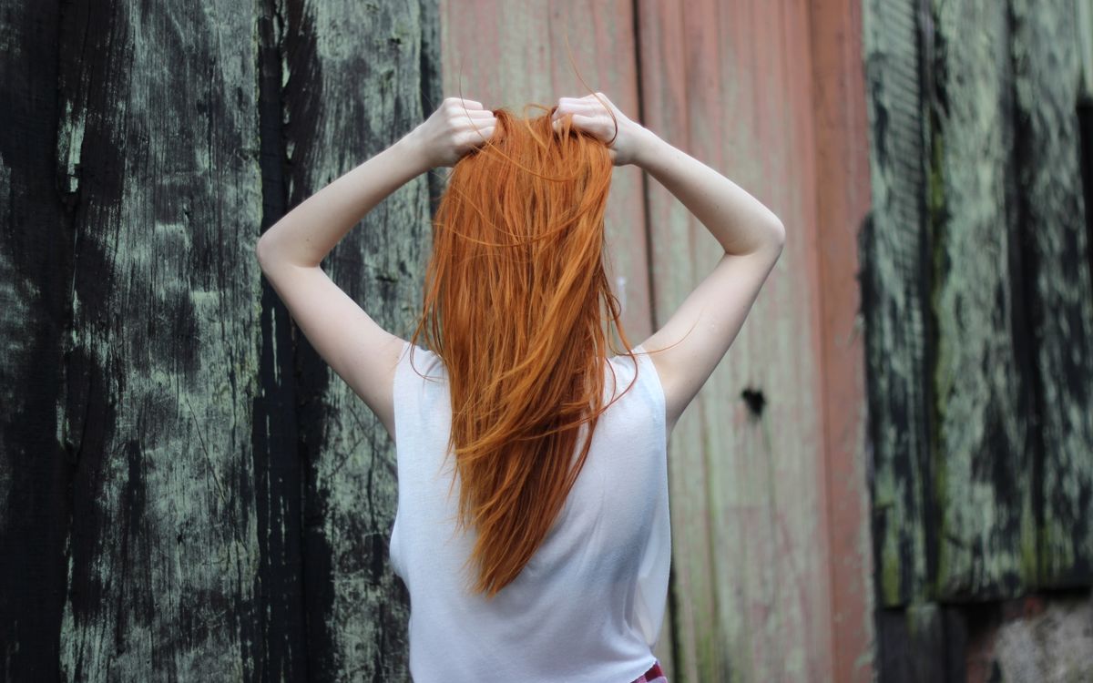 A red-haired woman poses with her back to the camera, outdoors against a rustic wooden backdrop.