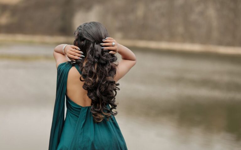 Back view of a woman in a flowing dress with curly hair by a serene lakeside setting.