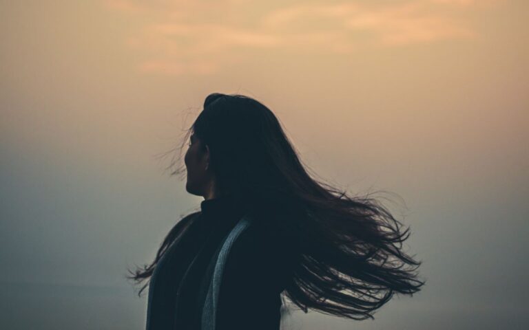 Side view of unrecognizable female shaking long hair while standing on shore near foggy sea on evening time in nature