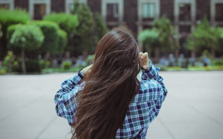 Rear view of a young woman with long hair in a checkered shirt adjusting her hair in an urban park setting.