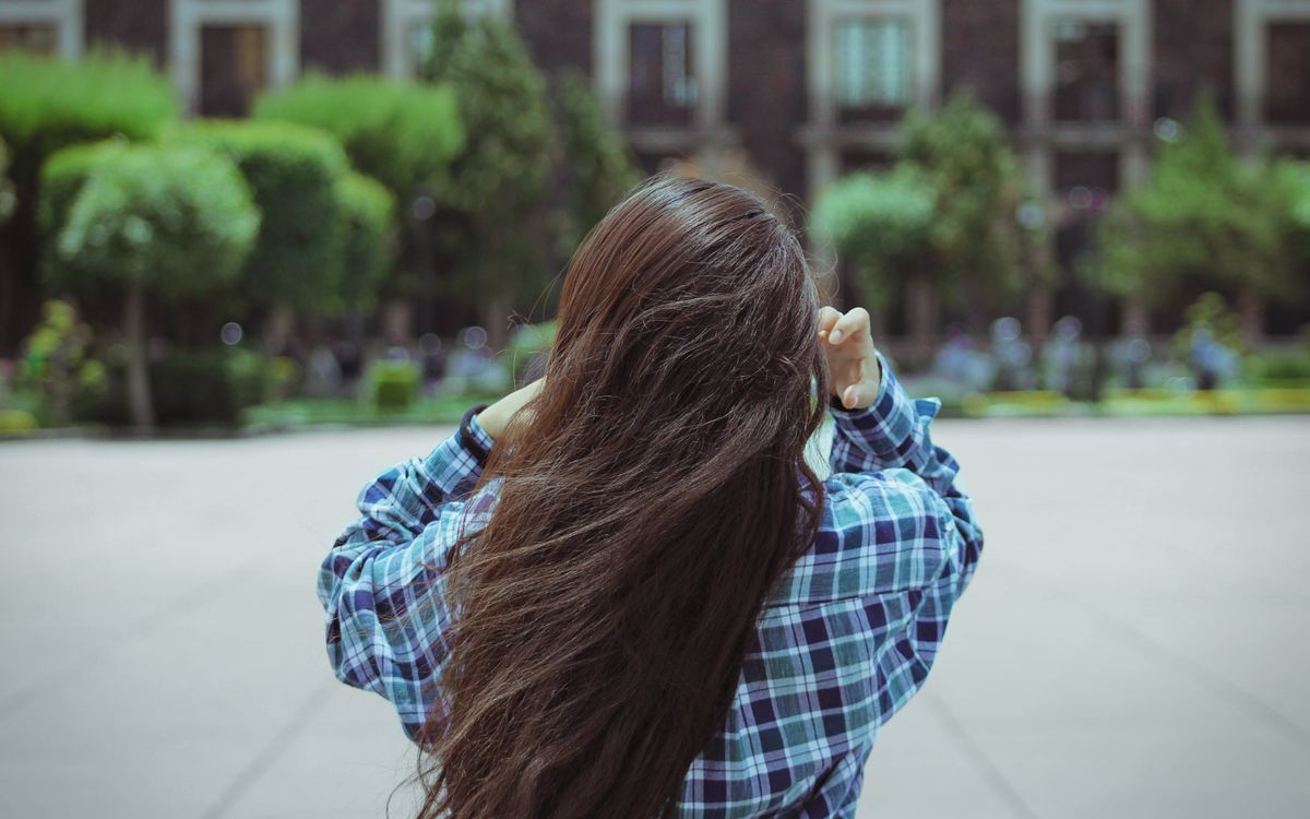 Rear view of a young woman with long hair in a checkered shirt adjusting her hair in an urban park setting.