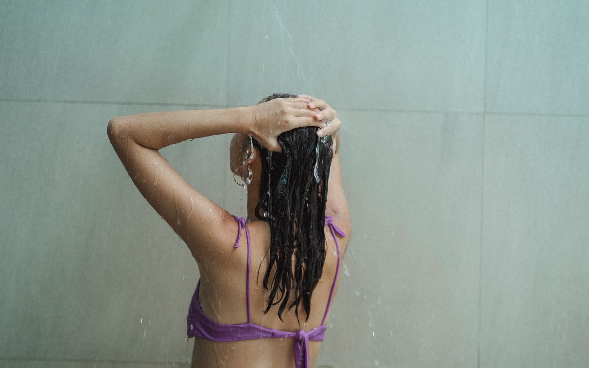 Back view of anonymous slim female wearing earrings and swimsuit touching hair while enjoying shower in bathroom