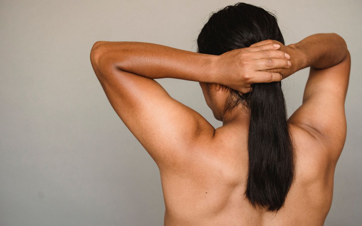 Back view anonymous male with naked torso adjusting long dark hair against gray wall in studio during photo session